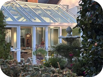 Ein  heller  Wintergarten  mit  Glasdach  zeigt  eine  gemütliche  Einrichtung  und  eine  Standuhr