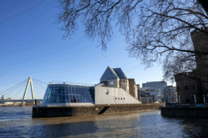 Modernes Gebäude mit Glasfront am Fluss vor blauen Himmel und Brücke im Hintergrund